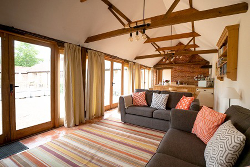 Internal view of cottage lounge area with striped rug, floor to ceiling windows and exposed beams and kitchen area at the end of the room